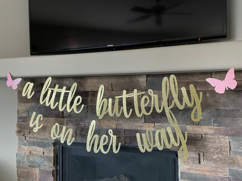 May include: A gold glitter banner with the phrase "a little butterfly is on her way" in cursive script. Pink butterfly cutouts are attached to the banner and placed on either side. The banner is strung across a stone fireplace.