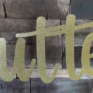 May include: Gold glitter cursive banner spelling out the word "Butter". The banner is strung on a thin, clear string and is set against a textured, grey brick wall. Ideal for a party or event.