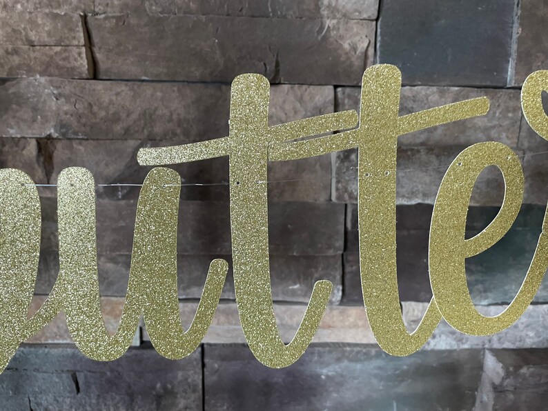 May include: Gold glitter cursive banner spelling out the word "Butter". The banner is strung on a thin, clear string and is set against a textured, grey brick wall. Ideal for a party or event.