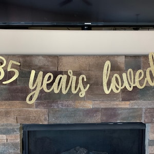 May include: A gold glitter banner that reads "85 years loved" is displayed against a stone fireplace. The banner is a celebratory decoration, perfect for an 85th birthday or anniversary.
