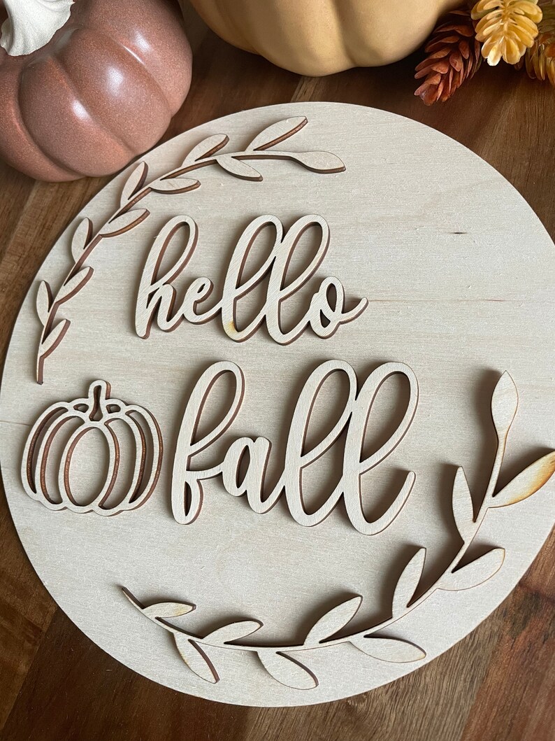 May include: A wooden round sign with the words "hello fall" in a cursive font. The sign is decorated with a pumpkin and leaves.