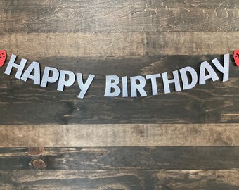 Może przedstawiać: Srebrny baner Happy Birthday z błyszczącymi literami i czerwonymi akcentami w postaci kontrolerów do gier. Baner jest zawieszony na ciemnym drewnianym tle.