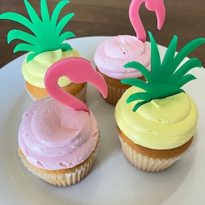 May include: Three cupcakes decorated with pink and yellow frosting and topped with flamingo and pineapple themed decorations.