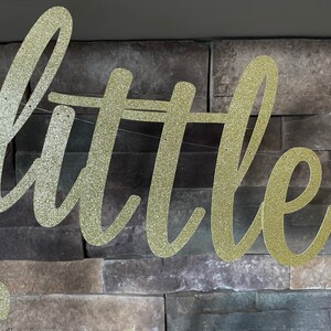 May include: A gold glitter banner spelling out the word "little" in a cursive font. The banner is strung on a thin white string and hangs against a textured, grey stone wall. The banner is likely for a party or celebration.