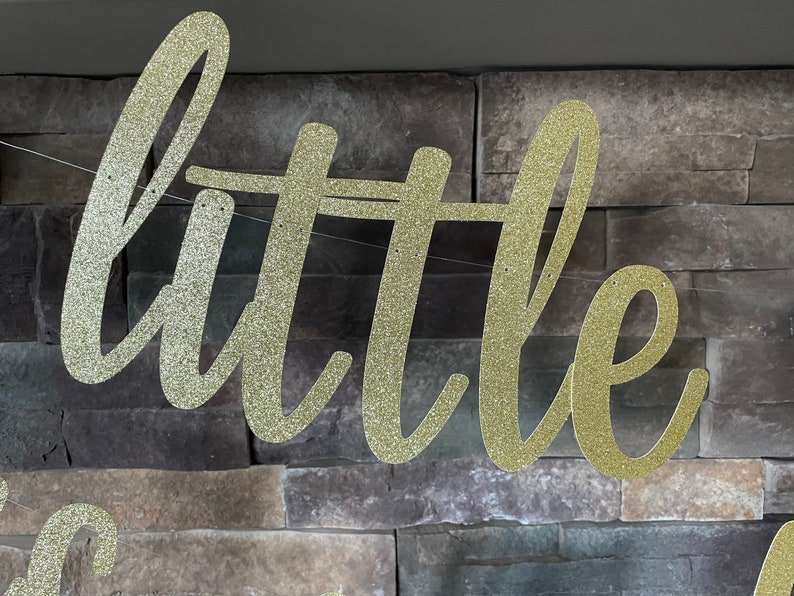 May include: A gold glitter banner spelling out the word "little" in a cursive font. The banner is strung on a thin white string and hangs against a textured, grey stone wall. The banner is likely for a party or celebration.