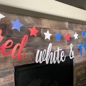 May include: A patriotic banner with red, white, and blue glitter stars and the words "red white & TWO". The banner is strung across a stone fireplace, perfect for a second birthday celebration.