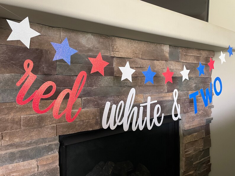 May include: A patriotic banner with red, white, and blue glitter stars and the words "red white & TWO". The banner is strung across a stone fireplace, perfect for a second birthday celebration.