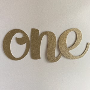 May include: Gold glitter "one" cutout. The word is in a cursive font. The cutout is likely for a birthday party or other celebration. The background is white.