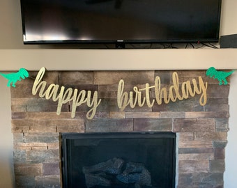 May include: A gold glitter "happy birthday" banner with green glitter dinosaur accents. The banner is displayed above a fireplace with a stone facade, creating a festive and celebratory atmosphere.