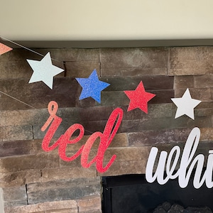 May include: A patriotic banner with red, white, and blue glittery star shapes strung together. The word "red" is in a cursive font, and the word "white" is also visible. The banner is displayed against a stone fireplace.