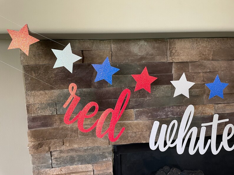 May include: A patriotic banner with red, white, and blue glittery star shapes strung together. The word "red" is in a cursive font, and the word "white" is also visible. The banner is displayed against a stone fireplace.