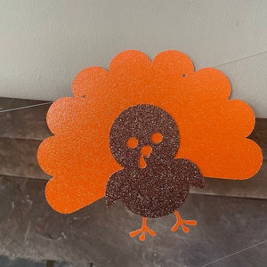 May include: Thanksgiving banner featuring a glittery turkey cutout. The turkey has a brown body and orange tail feathers. The banner is strung on a thin white string, perfect for holiday decorations.