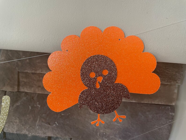 May include: Thanksgiving banner featuring a glittery turkey cutout. The turkey has a brown body and orange tail feathers. The banner is strung on a thin white string, perfect for holiday decorations.