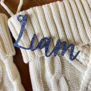 May include: White knitted stocking with a blue glitter name tag that says "Liam".