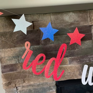 May include: A patriotic banner with glittery star cutouts in red, white, and blue, strung together. The word "red" is displayed in a cursive font. The banner is set against a stone fireplace.