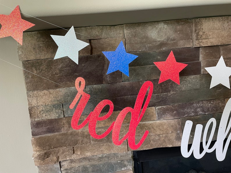 May include: A patriotic banner with glittery star cutouts in red, white, and blue, strung together. The word "red" is displayed in a cursive font. The banner is set against a stone fireplace.