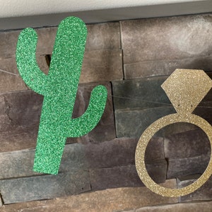 K&ouml;nnte beinhalten: Eine Girlande mit einem glitzernden gr&uuml;nen Kaktus und einem goldenen Verlobungsring. Der Kaktus ist gr&uuml;n und der Ring hat die Form eines Diamanten. Der Hintergrund ist eine Steinmauer, an der die Dekorationen h&auml;ngen.