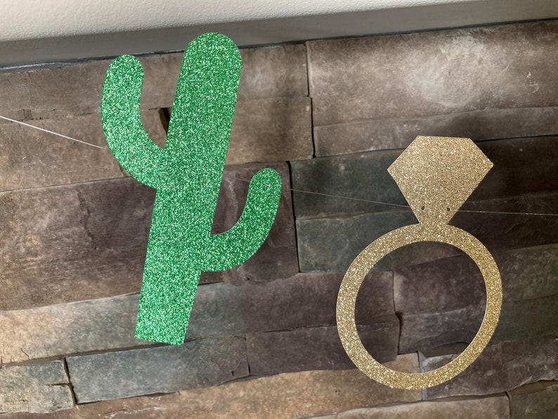 K&ouml;nnte beinhalten: Eine Girlande mit einem glitzernden gr&uuml;nen Kaktus und einem goldenen Verlobungsring. Der Kaktus ist gr&uuml;n und der Ring hat die Form eines Diamanten. Der Hintergrund ist eine Steinmauer, an der die Dekorationen h&auml;ngen.