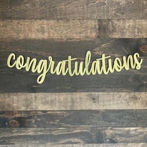 May include: A gold glitter banner with the word "congratulations" in a cursive font. The banner is displayed against a dark brown and gray wooden background. The banner is a decorative item for celebrations.