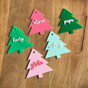 May include: A collection of colorful Christmas tree-shaped name tags. The tags are in shades of pink, green, and blue-green, each with a different name in white script. The tags are made of acrylic and have a small hole at the top for hanging.