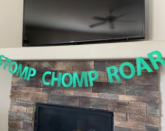 May include: A green glitter banner with the words "STOMP CHOMP ROAR" strung across. The banner is displayed in front of a stone fireplace and below a television.