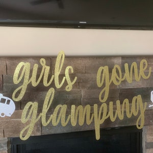 May include: A banner with the words "girls gone glamping" in gold glitter script. Two white camper trailer cutouts flank the text. The banner is strung across a stone fireplace.