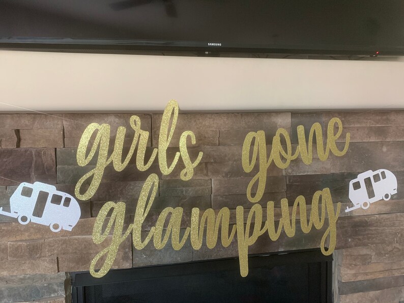 May include: A banner with the words "girls gone glamping" in gold glitter script. Two white camper trailer cutouts flank the text. The banner is strung across a stone fireplace.