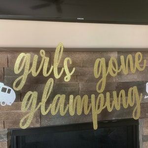 May include: A gold glitter banner with the words "girls gone glamping" in cursive script. Two white and silver camper trailer cutouts flank the text. The banner is strung against a stone wall.