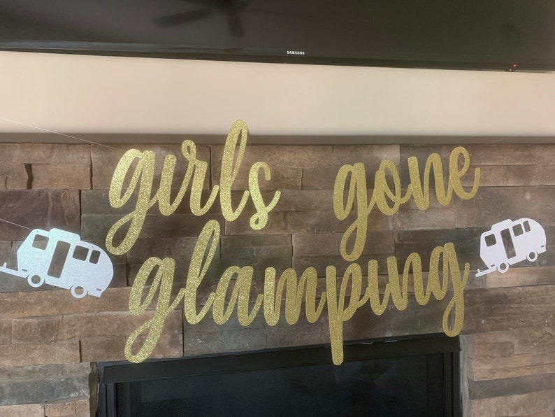 May include: A gold glitter banner with the words "girls gone glamping" in cursive script. Two white and silver camper trailer cutouts flank the text. The banner is strung against a stone wall.