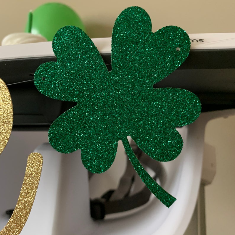 May include: A green glitter shamrock cutout with a hole in the top for hanging. The shamrock is a popular symbol of Irish culture and is often used in decorations for St. Patrick's Day.