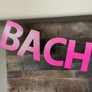 May include: A pink glitter banner spelling out the word "BACH" is strung across a room. The letters are large and bold, with a sparkly texture. The banner is likely for a bachelorette party or similar celebration.