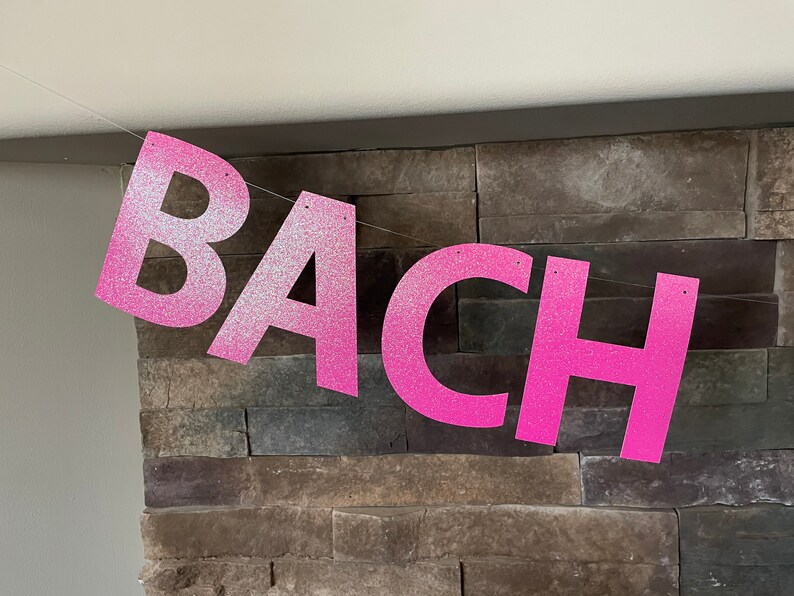 May include: A pink glitter banner spelling out the word "BACH" is strung across a room. The letters are large and bold, with a sparkly texture. The banner is likely for a bachelorette party or similar celebration.