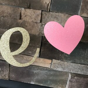 May include: A gold glitter letter "e" and a pink glitter heart are suspended against a textured brick wall. The letter is part of a word, possibly "love" or "one", creating a festive and decorative element.