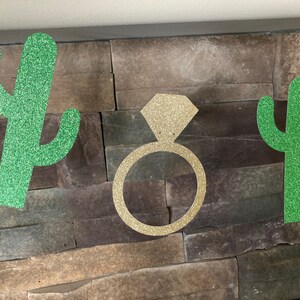 K&ouml;nnte beinhalten: Ein glitzerndes Banner mit einem goldenen Diamantring und zwei gr&uuml;nen Glitzer-Kaktus-Ausschnitten. Das Banner ist vor einer strukturierten Steinmauer aufgereiht, passend f&uuml;r eine Brautparty oder Verlobungsfeier.