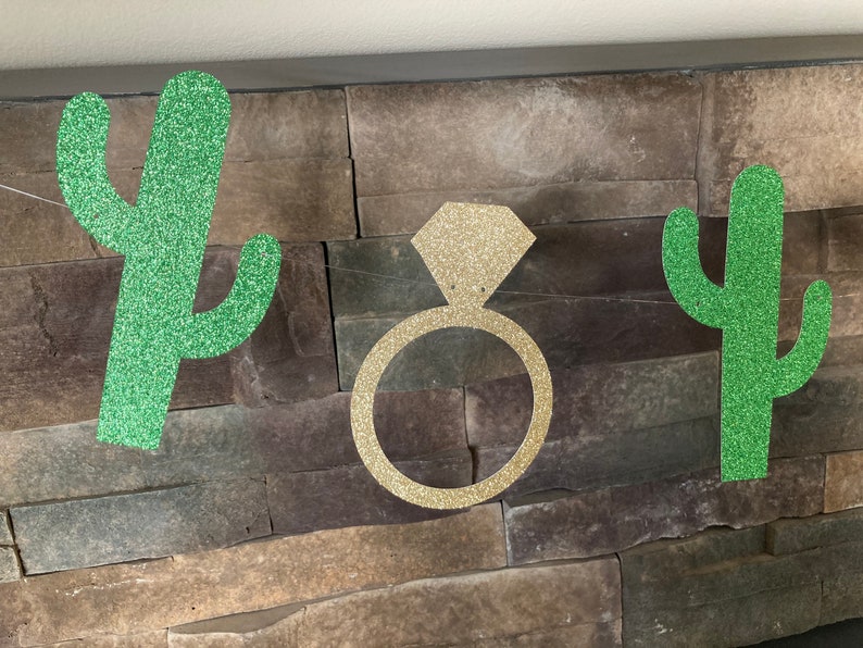 K&ouml;nnte beinhalten: Ein glitzerndes Banner mit einem goldenen Diamantring und zwei gr&uuml;nen Glitzer-Kaktus-Ausschnitten. Das Banner ist vor einer strukturierten Steinmauer aufgereiht, passend f&uuml;r eine Brautparty oder Verlobungsfeier.