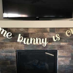 May include: A gold glitter banner that reads "some bunny is one" is displayed above a fireplace. The banner is made of individual letters and is designed for a first birthday celebration. The banner is hanging in front of a stone fireplace.