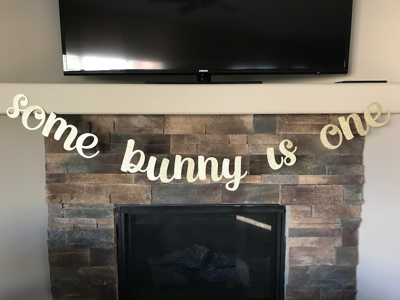May include: A gold glitter banner that reads "some bunny is one" is displayed above a fireplace. The banner is made of individual letters and is designed for a first birthday celebration. The banner is hanging in front of a stone fireplace.