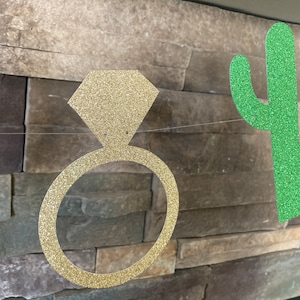 K&ouml;nnte beinhalten: Eine Girlande mit einem goldenen, glitzernden Verlobungsring und einem gr&uuml;nen Kaktus-Ausschnitt, auf einem Draht aufgereiht. Die Dekorationen sind vor einer strukturierten Steinmauer angebracht, passend f&uuml;r eine Brautparty oder Verlobungsfeier.