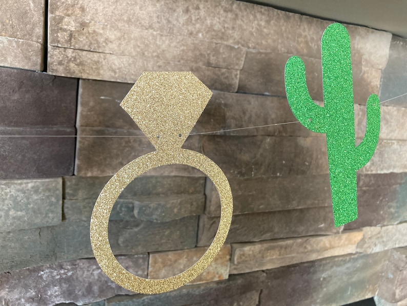K&ouml;nnte beinhalten: Eine Girlande mit einem goldenen, glitzernden Verlobungsring und einem gr&uuml;nen Kaktus-Ausschnitt, auf einem Draht aufgereiht. Die Dekorationen sind vor einer strukturierten Steinmauer angebracht, passend f&uuml;r eine Brautparty oder Verlobungsfeier.