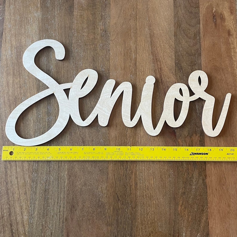 May include: A wooden cutout of the word "Senior" in a cursive font. The cutout is approximately 18 inches long and is shown on a wooden surface with a ruler for scale.