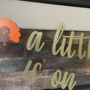 May include: Thanksgiving-themed banner with gold glitter lettering that reads "a little is on the". A glittery turkey decoration is on the left. The banner is strung across a stone wall.