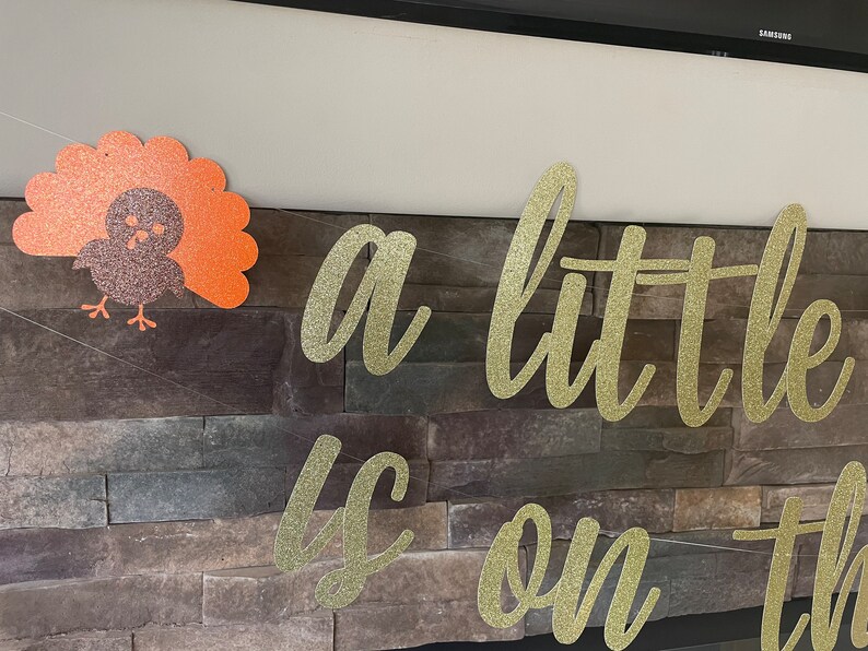 May include: Thanksgiving-themed banner with gold glitter lettering that reads "a little is on the". A glittery turkey decoration is on the left. The banner is strung across a stone wall.