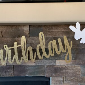 May include: A gold glitter "birthday" banner with cursive lettering is displayed against a stone fireplace. A white glitter bunny cutout hangs to the right, adding a festive touch. Perfect for a birthday celebration.