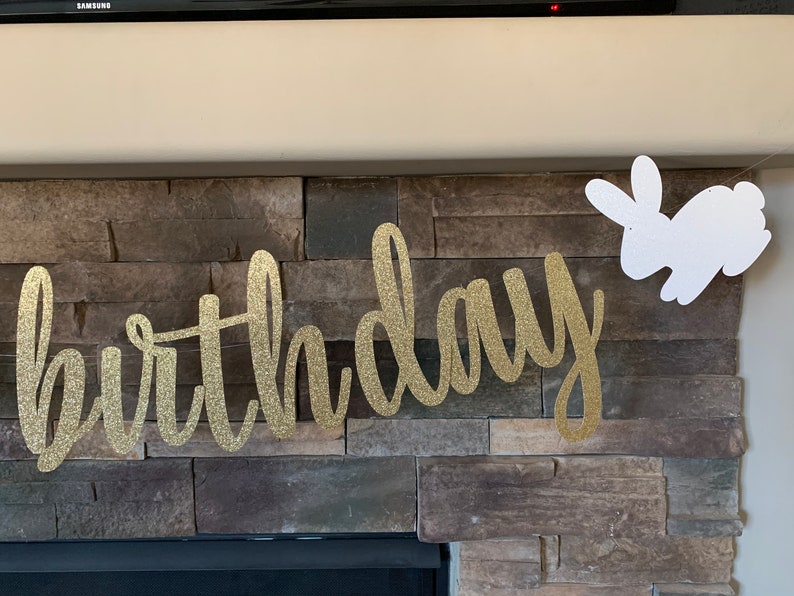May include: A gold glitter "birthday" banner with cursive lettering is displayed against a stone fireplace. A white glitter bunny cutout hangs to the right, adding a festive touch. Perfect for a birthday celebration.