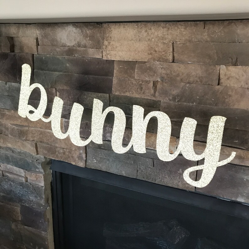 May include: A glittery "bunny" banner in a cursive font hangs in front of a stone fireplace. The banner is strung on a thin, clear string. The word "bunny" is spelled out in a sparkly, light-colored material.