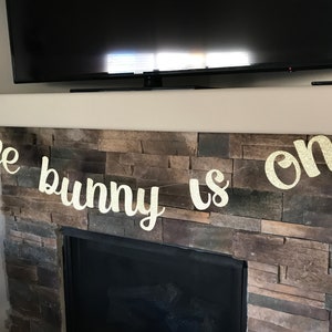 May include: Glittery gold banner with the phrase "some bunny is one" in cursive script. The banner is strung across a fireplace mantel, perfect for a first birthday celebration. The letters are cut out and have a sparkly finish.