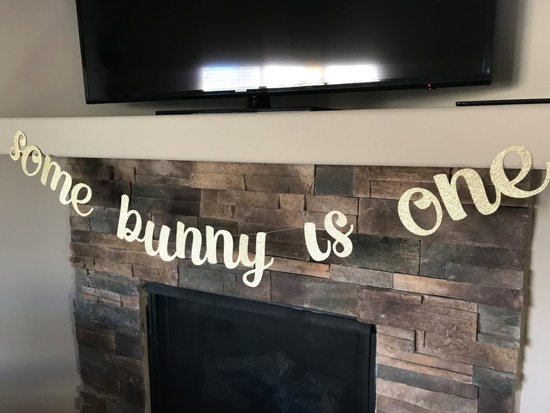 May include: Glittery gold banner with the phrase "some bunny is one" in cursive script. The banner is strung across a fireplace mantel, perfect for a first birthday celebration. The letters are cut out and have a sparkly finish.