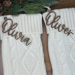 May include: Two white knitted stockings with wooden name tags. The stockings have a cable knit pattern. The name tags are in cursive and read "Olivia" and "Oliver".