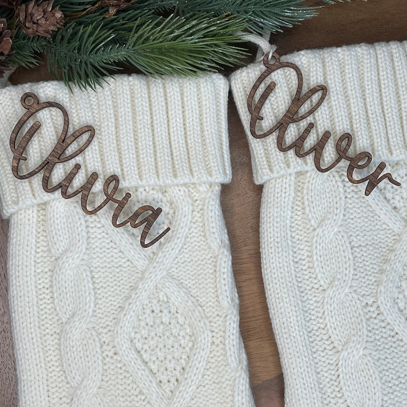 May include: Two white knitted stockings with wooden name tags. The stockings have a cable knit pattern. The name tags are in cursive and read "Olivia" and "Oliver".
