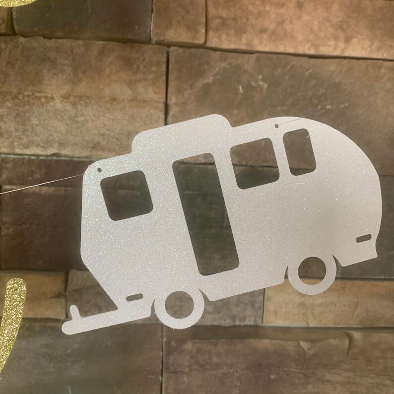 May include: A glittery silver camper banner with a rectangular window, two round wheels, and a hitch. The banner is strung on a thin, clear string. The camper is cut out of a sparkly material, perfect for a camping-themed party.